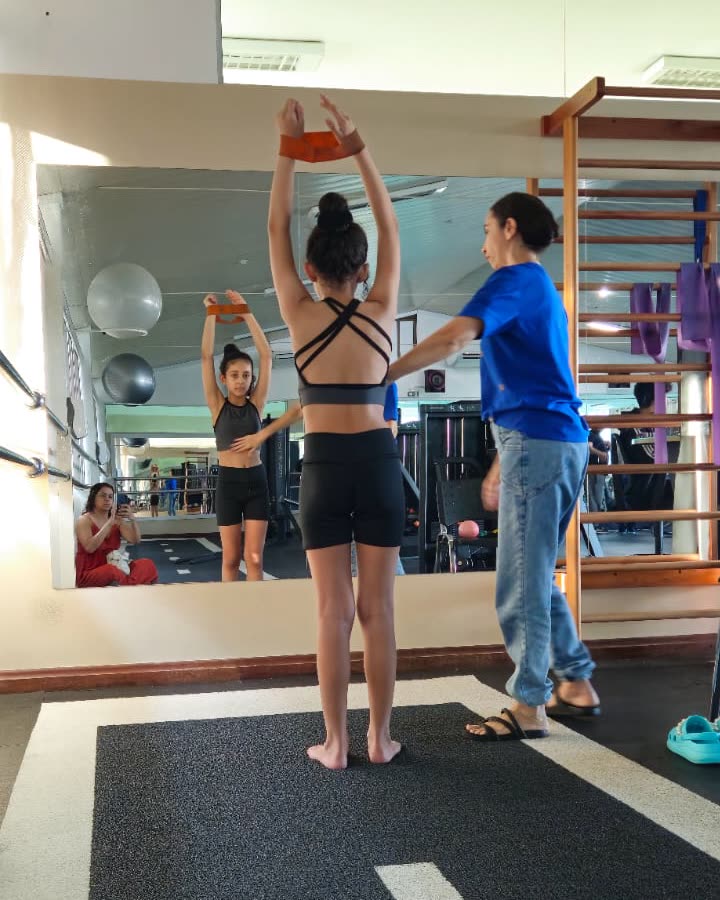 Giovana ajustando a postura de uma paciente durante um exercício com faixa acima da cabeça